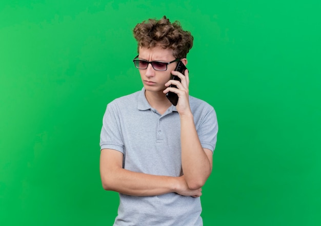 Hombre joven con gafas negras con camisa polo gris mirando a un lado confundido mientras habla por teléfono móvil sobre verde