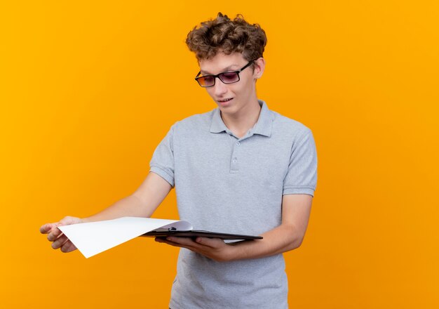 Hombre joven con gafas negras con camisa de polo gris con portapapeles con páginas en blanco mirándolo con una sonrisa en la cara sobre naranja