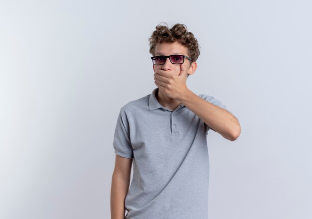 Hombre joven de gafas negras con camisa de polo gris que cubre la boca con la mano que se sorprende de pie sobre la pared blanca