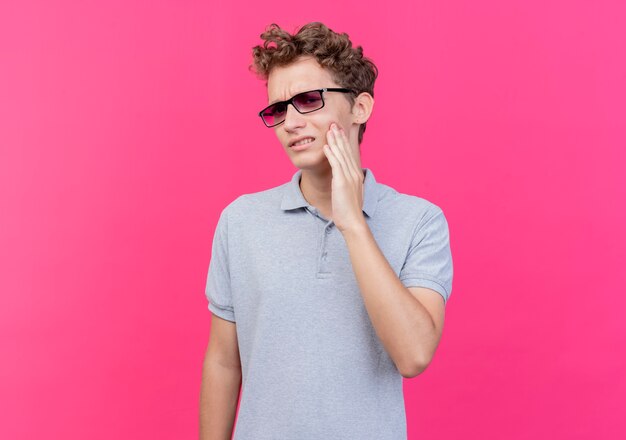 Hombre joven con gafas negras con camisa de polo gris que parece enfermo tocando su mejilla con dolor de muelas de pie sobre la pared rosa