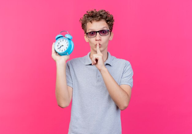 Hombre joven con gafas negras con camisa de polo gris con reloj despertador haciendo gesto de silencio con el dedo en los labios de pie sobre la pared rosa