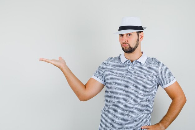 Hombre joven gesticulando como dando la bienvenida o mostrando algo en camiseta rayada, sombrero y mirando atenta, vista frontal.