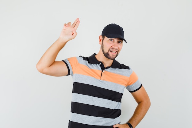 Hombre joven gesticulando con la mano y los dedos en camiseta, gorra y mirando confiado. vista frontal.