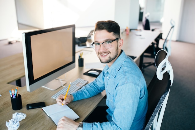 Hombre joven en glassess está trabajando en su lugar de trabajo en la oficina. Viste camisa azul. Está escribiendo en un cuaderno y sonriendo a la cámara.