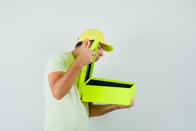 Hombre joven con gorra de camiseta mirando en caja de regalo y mirando curioso