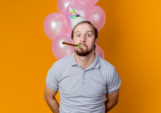 Hombre joven con gorro de fiesta celebrando la fiesta de cumpleaños con globos sorprendido de pie sobre la pared naranja