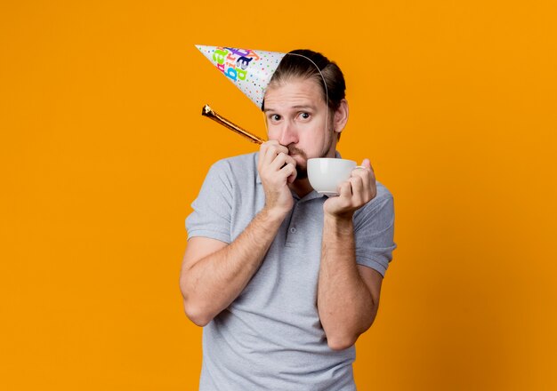 Hombre joven con gorro de fiesta celebrando la fiesta de cumpleaños con gorro de café mirando sorprendido y confundido de pie sobre la pared naranja