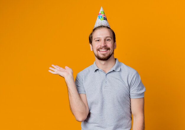 Hombre joven con gorro de fiesta celebrando la fiesta de cumpleaños presentando algo con el brazo feliz y alegre sonriendo ampliamente de pie sobre la pared naranja