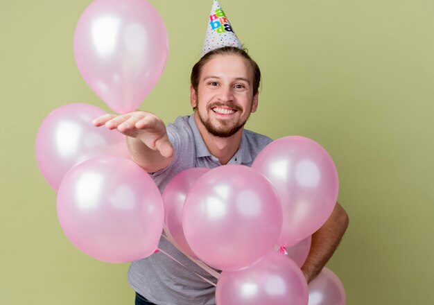 Hombre joven con gorro de fiesta celebrando la fiesta de cumpleaños sosteniendo globos alegre y emocionado sonriendo alegremente de pie sobre la pared de luz