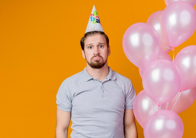 Hombre joven con gorro de fiesta celebrando la fiesta de cumpleaños sosteniendo globos con expresión triste en la cara de pie sobre la pared naranja