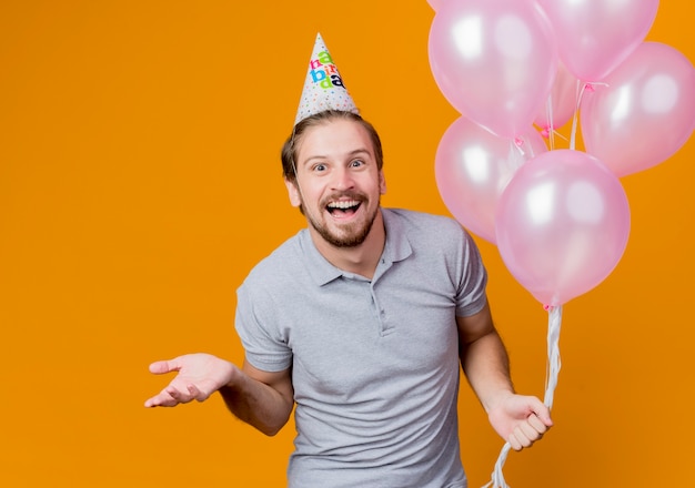 Hombre joven con gorro de fiesta celebrando la fiesta de cumpleaños sosteniendo globos sonriendo alegremente ingenio brazo de pie sobre la pared naranja