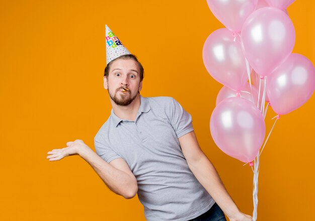 Hombre joven con gorro de fiesta celebrando la fiesta de cumpleaños sosteniendo un montón de globos mirando sorprendido y asombrado de pie sobre la pared naranja