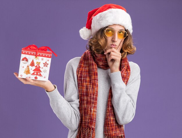 Hombre joven con gorro de navidad de santa y gafas amarillas con bufanda alrededor de su cuello sosteniendo un regalo de navidad mirando hacia arriba con expresión pensativa pensando de pie sobre la pared púrpura