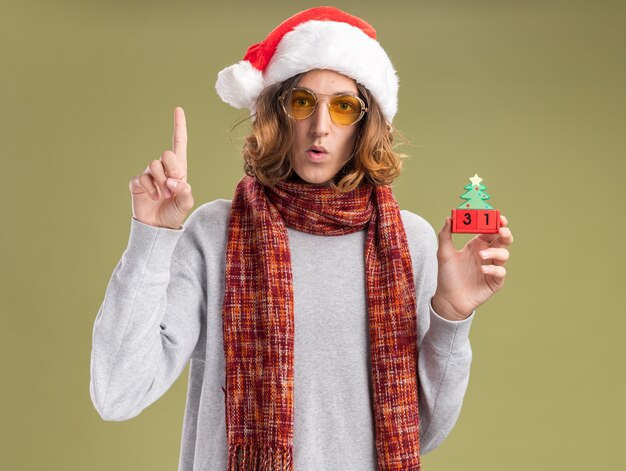 Hombre joven con gorro de navidad santa y gafas amarillas con bufanda caliente alrededor de su cuello sosteniendo cubos de juguete con fecha de año nuevo mostrando el dedo índice preocupado de pie sobre la pared verde