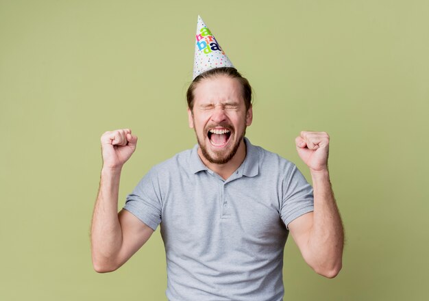 Hombre joven con gorro navideño celebrando la fiesta de cumpleaños feliz y emocionado sobre pared ligera