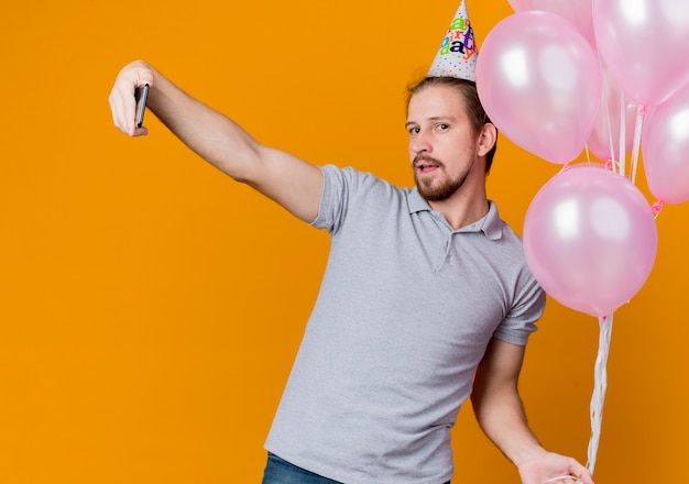 Hombre joven con gorro navideño celebrando la fiesta de cumpleaños sosteniendo un montón de globos haciendo selfie con smartphone parado sobre pared naranja