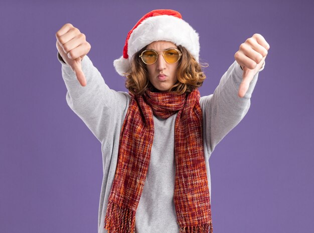 Hombre joven con gorro de Papá Noel de Navidad y gafas amarillas con bufanda alrededor de su cuello mirando a la cámara con el ceño fruncido serio mostrando los pulgares hacia abajo de pie sobre fondo púrpura