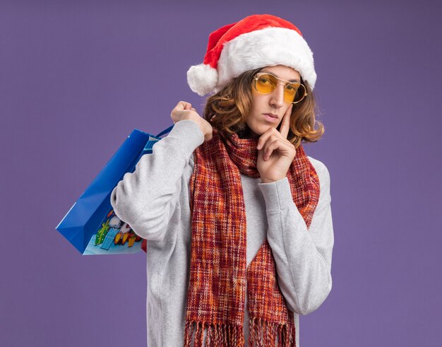 Hombre joven con gorro de Papá Noel de Navidad y gafas amarillas con bufanda alrededor de su cuello sosteniendo bolsas de papel de Navidad con regalos mirando a la cámara con cara seria de pie sobre fondo púrpura