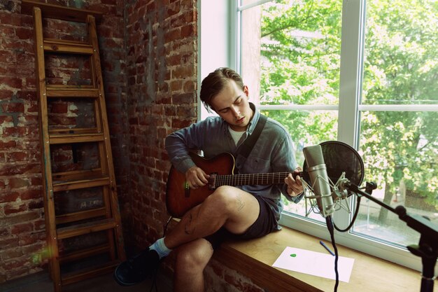 Hombre joven grabando un blog de videos musicales, una lección o una canción en casa, tocando la guitarra o haciendo un tutorial de transmisión por Internet mientras está sentado en el lugar de trabajo del loft o en casa. Concepto de afición, música, arte y creación.