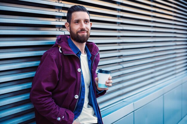 Hombre joven guapo con chaqueta de invierno púrpura bebiendo café. El tipo bebiendo café en la calle. Hombre sosteniendo taza de papel con americano o latte. Estilo de calle