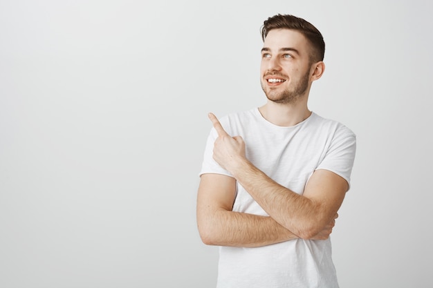 Foto gratuita hombre joven guapo complacido en camiseta blanca que señala el dedo en la esquina superior izquierda