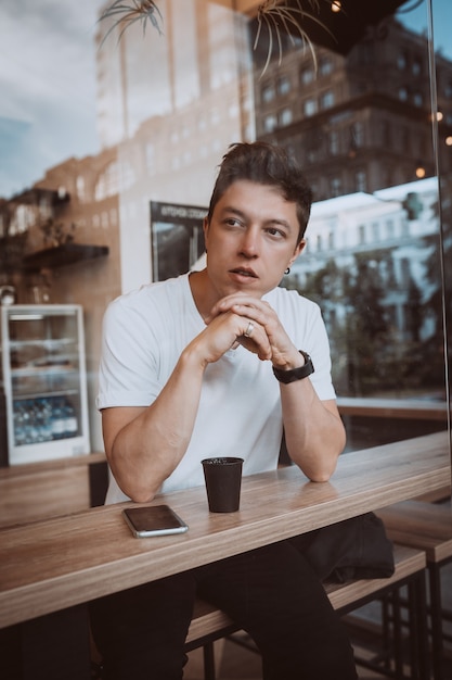 Hombre joven y guapo está bebiendo su café de la mañana en un café. Foto detrás de un vidrio
