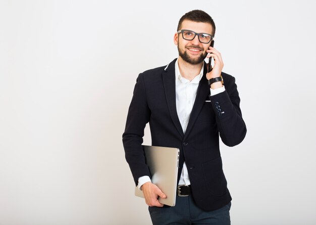 Hombre joven guapo con estilo hipster en chaqueta negra, estilo empresarial, camisa blanca, aislado, fondo blanco, sonriente, atractivo, confiado, sosteniendo la computadora portátil, hablando por teléfono inteligente