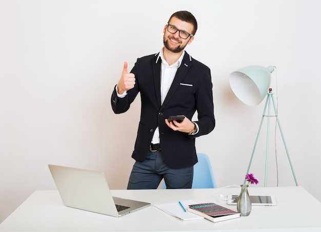 Hombre joven guapo con estilo hipster en chaqueta negra en la mesa de oficina, estilo empresarial, camisa blanca, aislado, trabajando en la computadora portátil, puesta en marcha, lugar de trabajo, sonriente, feliz, positivo,