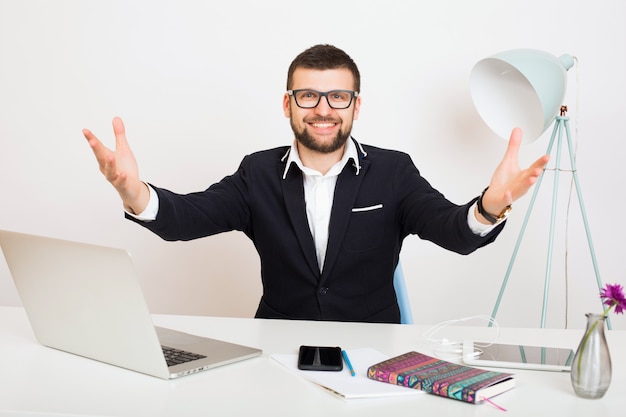 Hombre joven guapo con estilo hipster en chaqueta negra en la mesa de la oficina, estilo de negocios, camisa blanca, aislado, trabajando en la computadora portátil, puesta en marcha, lugar de trabajo, saludo de brazos abiertos, sonriente, feliz, positivo