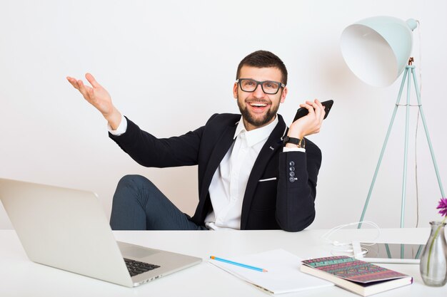 Hombre joven guapo con estilo hipster en chaqueta negra sentado en la mesa de la oficina, estilo de negocios, camisa blanca, aislado, trabajando, computadora portátil, puesta en marcha, lugar de trabajo, hablando por teléfono inteligente, sonriendo, positivo