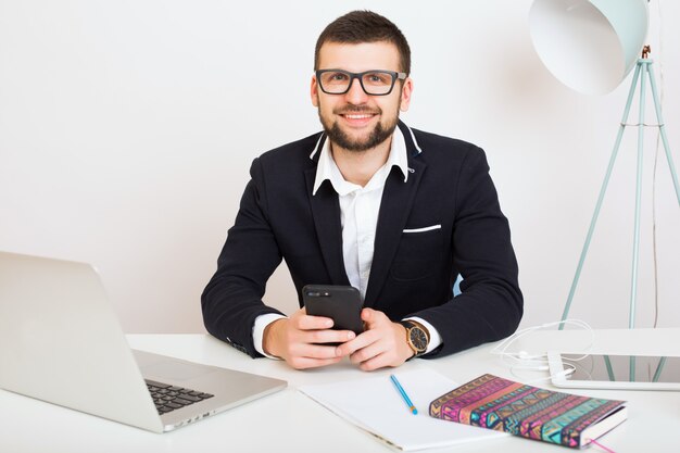 Hombre joven guapo con estilo hipster en chaqueta negra sentado en la mesa de la oficina, estilo de negocios, camisa blanca, aislado, trabajando, computadora portátil, puesta en marcha, lugar de trabajo, hablando por teléfono inteligente, sonriendo, positivo