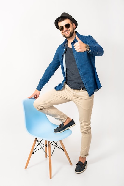 Hombre joven guapo hipster, fondo de estudio blanco aislado, traje elegante, camisa de mezclilla, pantalones, sombrero, gafas de sol, de pie en la silla, alegre, feliz, sonriente, positivo, éxito, alegre, pulgar hacia arriba