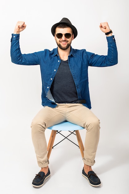 Hombre joven guapo hipster, fondo de estudio blanco aislado, traje elegante, camisa de mezclilla, pantalones, sombrero, gafas de sol, sentado en una silla, éxito, ganar, emocional, feliz, sonriente