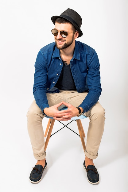 Hombre joven guapo hipster, fondo de estudio blanco aislado, traje elegante, camisa de mezclilla, pantalones, sombrero, gafas de sol, sentado en una silla, manos juntas, pose, sonriente, feliz, positivo