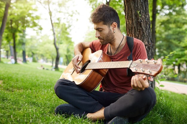 Hombre joven guapo pensativo tocando la guitarra en el parque, apoyado en un árbol y sentarse en el césped