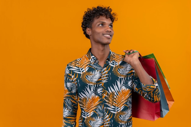 Hombre joven guapo de piel oscura con cabello rizado en hojas de camisa estampada sonriendo sosteniendo bolsas de compras mientras está de pie sobre un fondo naranja