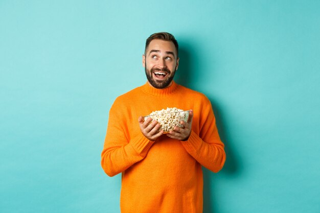 Hombre joven guapo en suéter naranja, mirando pensativo en la esquina superior izquierda, sosteniendo palomitas de maíz