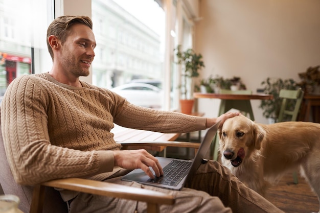 Foto gratuita hombre joven guapo trabajando en una cafetería con un perro sentado en una silla y usando una computadora portátil acariciando su dorada