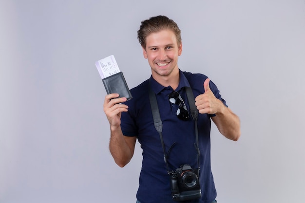 Hombre joven guapo viajero con cámara sosteniendo boletos de avión mirando a cámara con sonrisa en la cara feliz y positivo mostrando los pulgares para arriba sobre fondo blanco