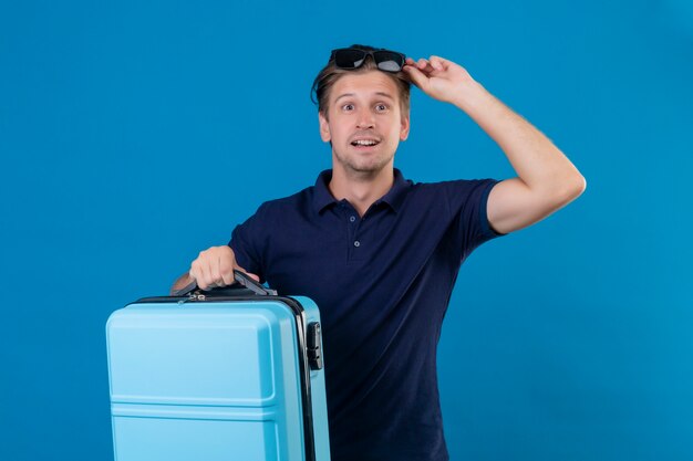 Hombre joven guapo viajero con maleta mirando salido y feliz listo para viajar de pie sobre fondo azul.