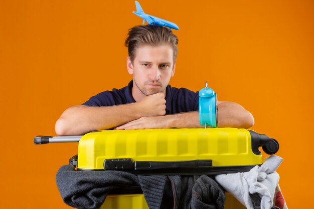 Hombre joven guapo viajero de pie con la maleta llena de ropa con reloj despertador y con avión de juguete en la cabeza con aspecto cansado y aburrido con expresión triste en la cara sobre fondo naranja