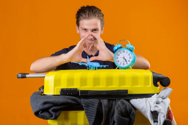 Hombre joven guapo viajero de pie con la maleta llena de ropa con reloj despertador y avión de juguete haciendo gesto de corazón con las manos mirando positivo y feliz sonriendo sobre fondo naranja