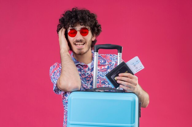 Hombre joven guapo viajero rizado complacido con gafas de sol sosteniendo boletos de avión, billetera poniendo las manos en la cabeza y en la maleta con los ojos cerrados en el espacio rosa aislado con espacio de copia