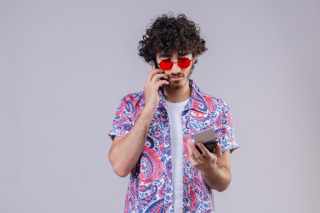 Hombre joven guapo viajero rizado con gafas de sol hablando por teléfono sosteniendo boletos de avión y billetera y mirándolos en una pared blanca aislada con espacio de copia