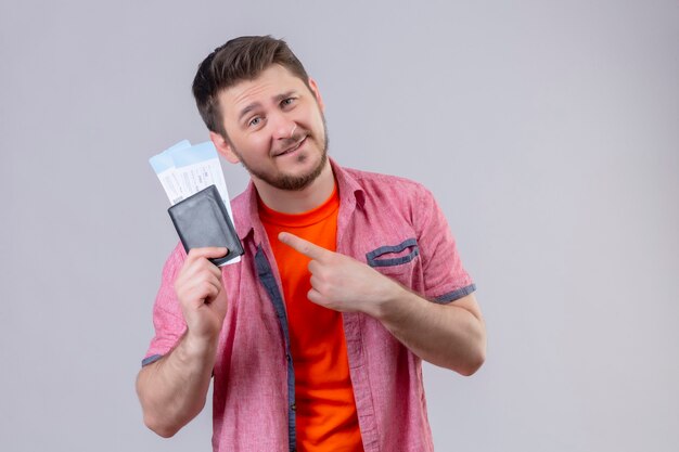 Hombre joven guapo viajero sosteniendo billetes de avión apuntando con el dedo a ellos sonriendo positivo y feliz de pie sobre la pared blanca