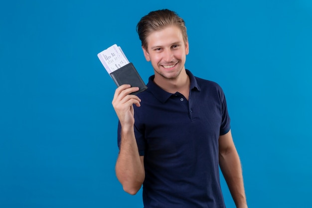 Hombre joven guapo viajero sosteniendo billetes de avión feliz y positivo mirando a cámara con gran sonrisa en la cara de pie sobre fondo azul