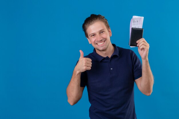 Hombre joven guapo viajero sosteniendo billetes de avión sonriendo alegremente con cara feliz mostrando los pulgares para arriba sobre fondo azul