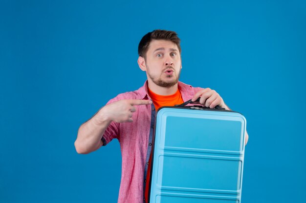 Hombre joven guapo viajero sosteniendo la maleta apuntando con el dedo hacia el lado con expresión confusa en la cara de pie sobre la pared azul
