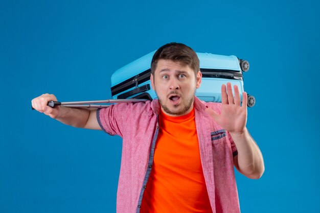 Hombre joven guapo viajero sosteniendo maleta haciendo señal de stop con la mano asustada de pie sobre la pared azul