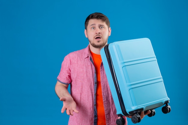 Foto gratuita hombre joven guapo viajero sosteniendo la maleta mirando confundido y muy ansioso de pie sobre la pared azul